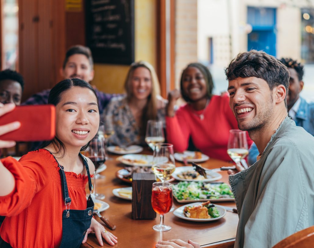 Asian woman taking a selfie at a multiethnic gathering of friends in a restaurant with a red cell phone