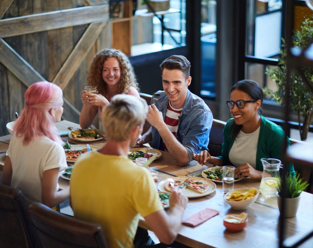 Happy guys and girls having talk by dinner while sitting by served table in cafe